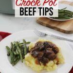 Crock pot beef tips over a bed of mashed potatoes accompanied by some green beans sit on a plate upon the kitchen counter, a full plate of green beans and the crock pot are in the background.