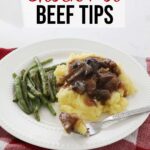 Crock pot beef tips over a bed of mashed potatoes accompanied by some green beans sit on a white plate on the kitchen counter, on a red towel.