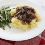 Crock pot beef tips over a bed of mashed potatoes accompanied by some green beans sit on a white plate on the kitchen counter, on a red towel and a fork in the foreground.