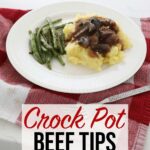 Crock pot beef tips over a bed of mashed potatoes accompanied by some green beans sit on a white plate on the kitchen counter, on a red towel, and a fork in the foreground.