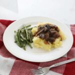 Crock pot beef tips over a bed of mashed potatoes accompanied by some green beans sit on a white plate on the kitchen counter, on a red towel with a fork In the foreground.