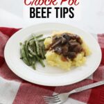 Crock pot beef tips over a bed of mashed potatoes accompanied by some green beans sit on a white plate on the kitchen counter, on a red towel with a fork In the foreground.
