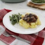 Crock pot beef tips over a bed of mashed potatoes accompanied by some green beans sit on a plate upon the kitchen counter, with a fork in the foreground and full plate of green beans in the background.