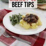 Crock pot beef tips over a bed of mashed potatoes accompanied by some green beans sit on a plate upon the kitchen counter, with a fork in the foreground and full plate of green beans in the background.