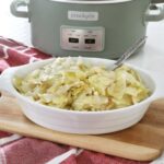 A large serving dish of crock pot cabbage sits on a wooden board, on a red kitchen towel with the crock pot in display behind it.