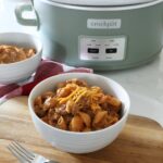 A white ceramic bowl full of hamburger helper rests on top of a wooden board on top of the kitchen counter, with a fork in the foreground and a crock pot along with another bowl in background.