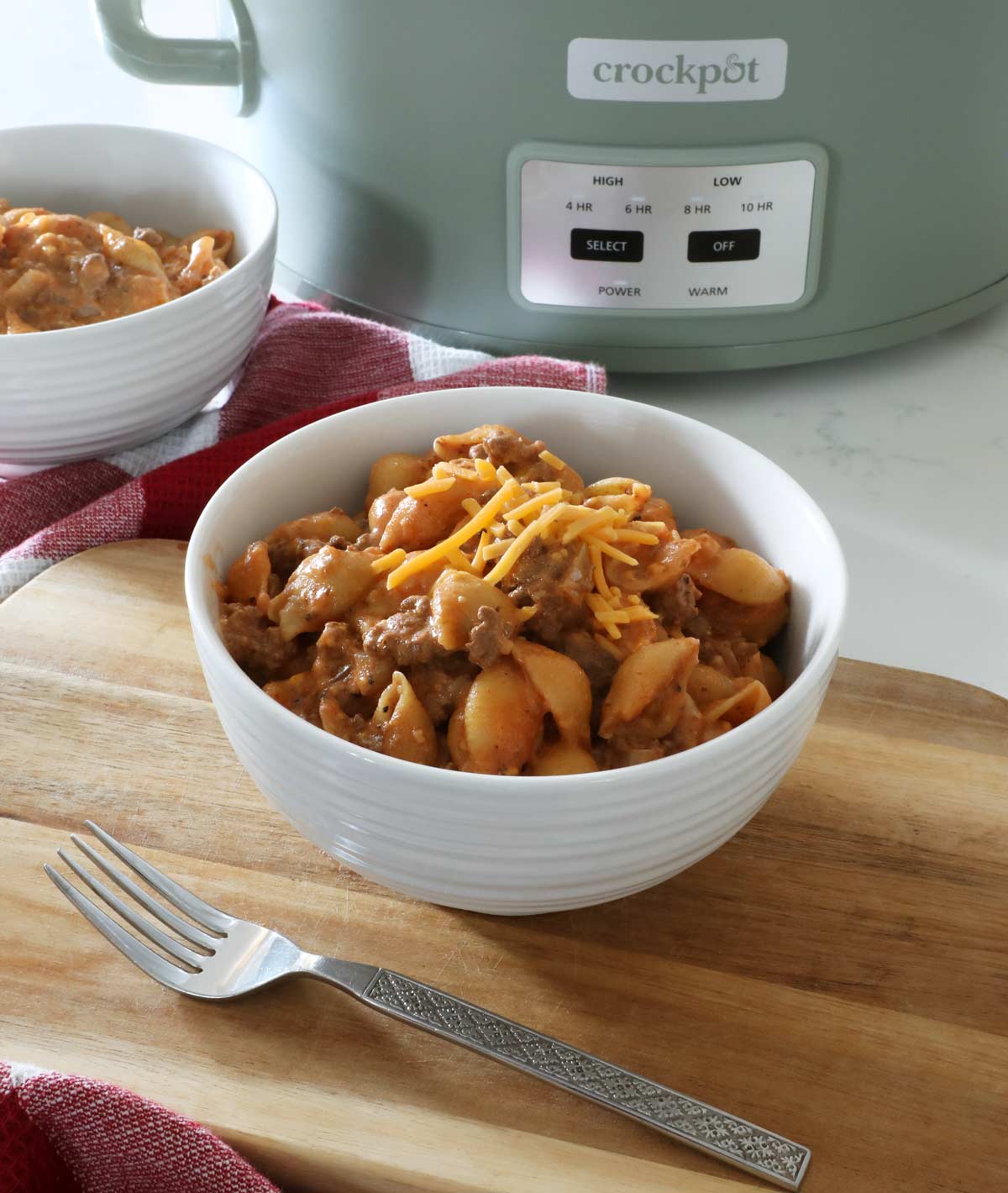 A white ceramic bowl full of hamburger helper rests on top of a wooden board and red towel on top of the kitchen counter, with a fork in the foreground and a crock pot along with another bowl in background.