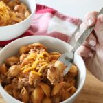 A white ceramic bowl full of hamburger helper rests on top of a wooden board and red towel on top of the kitchen counter, a person's hand is in frame using the fork to scoop the hamburger helper.