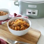 A white ceramic bowl full of hamburger helper rests on top of a wooden board on top of the kitchen counter, with a fork in the foreground and a crock pot along with another bowl in background.