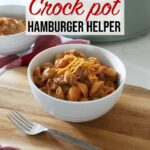 A white ceramic bowl full of hamburger helper rests on top of a wooden board and red towel on top of the kitchen counter, with a fork in the foreground and a crock pot along with another bowl in background.