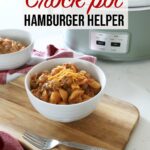 A white ceramic bowl full of hamburger helper rests on top of a wooden board on top of the kitchen counter, with a fork in the foreground and a crock pot along with another bowl in background.