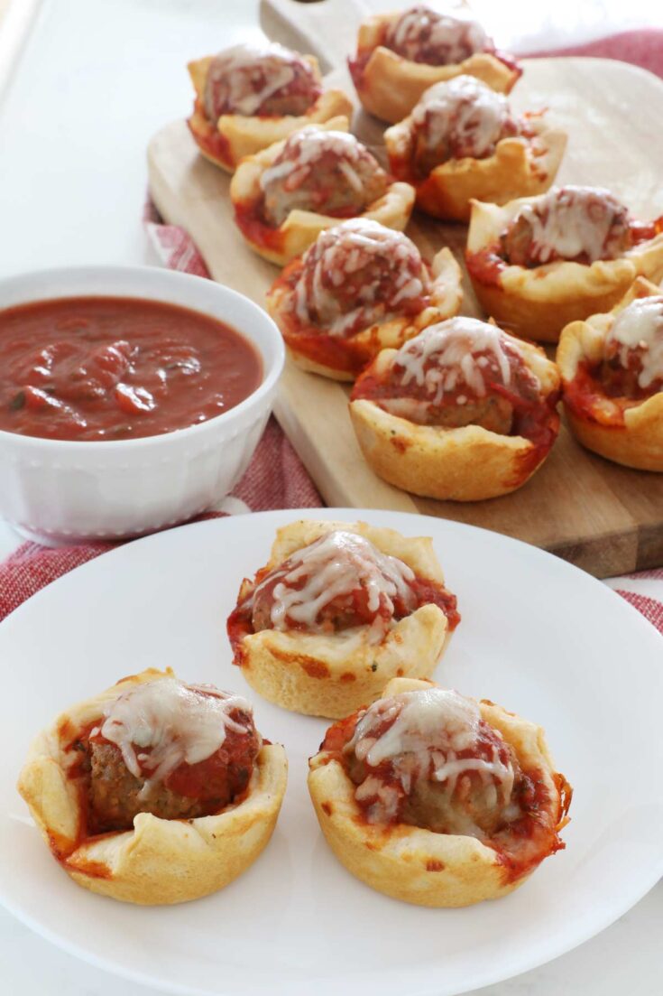 3 Individual Meatball Subs sit on a white plate on top of the kitchen counter, 8 more sit on a wooden board in the background, a side bowl of marinara sauce for dipping is to the left of everything.