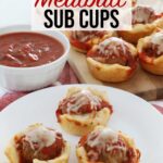 3 Meatball cups sit on a white plate on top of the kitchen counter, 8 more sit on a wooden board in the background, a side bowl of marinara sauce for dipping is to the left of everything.