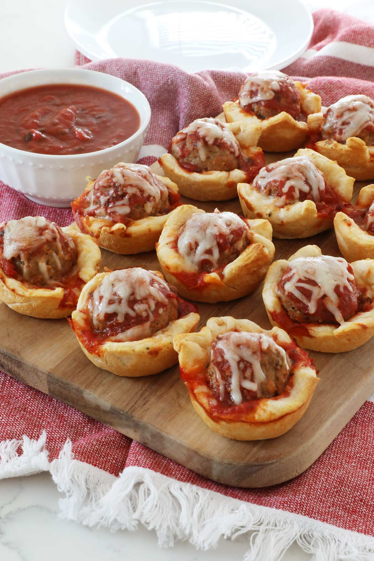 Meatball sub cups sit lined up neatly on a wooden board, on top of a red kitchen towel along with a bowl of marinara sauce for dipping and an empty white plate.