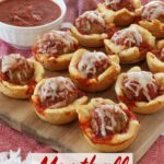 Meatball cups sit lined up neatly on a wooden board, on top of a red kitchen towel along with a bowl of marinara sauce for dipping and an empty white plate.