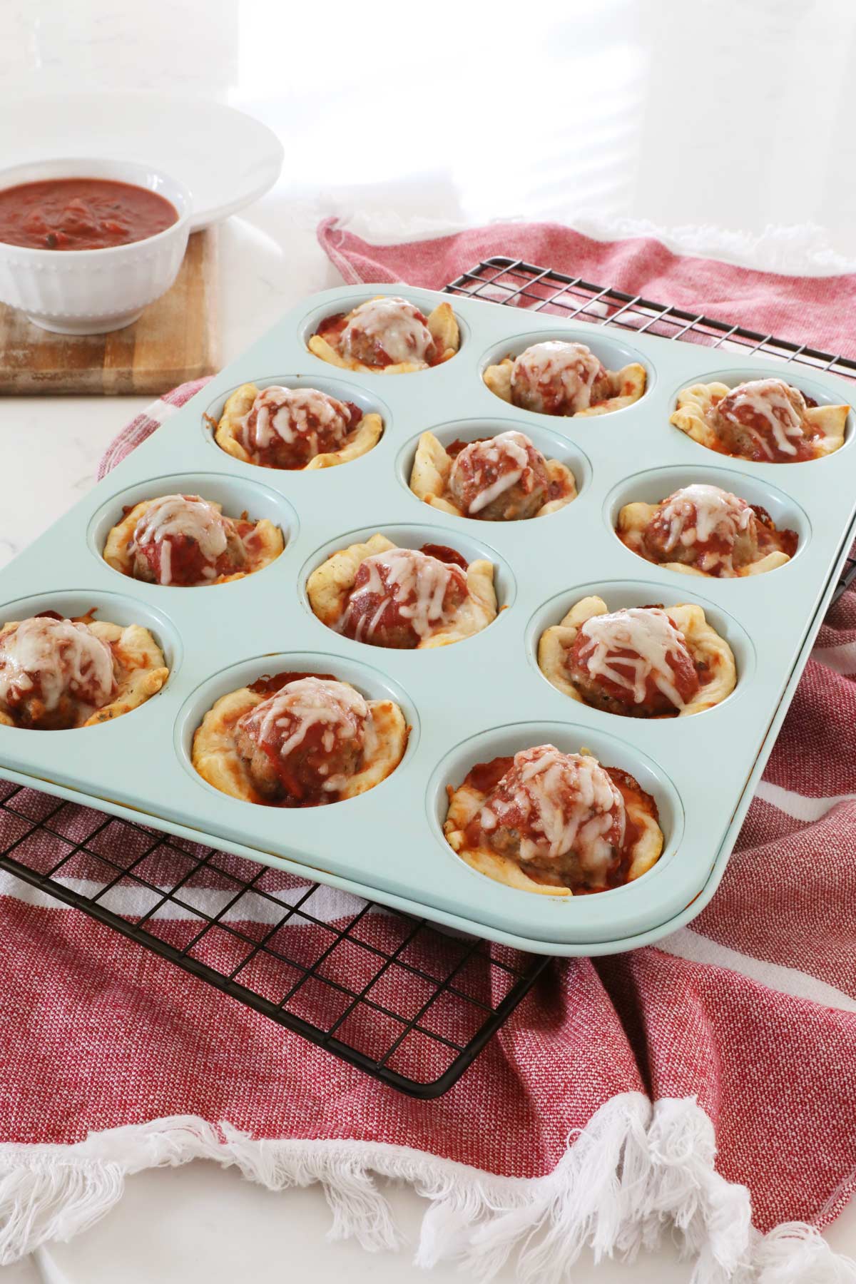 Meatball Sub cups sitting in a teal muffin tin on the kitchen counter, marinara dipping sauce sits in a bowl off the the side.