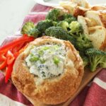 A close up of a bread bowl full of Knorr spinach dip sits on a wooden board along with some broccoli florets, sliced red bell pepper and more bread pieces, a cheese spreader sits on a white plate in the background on top of the kitchen counter.