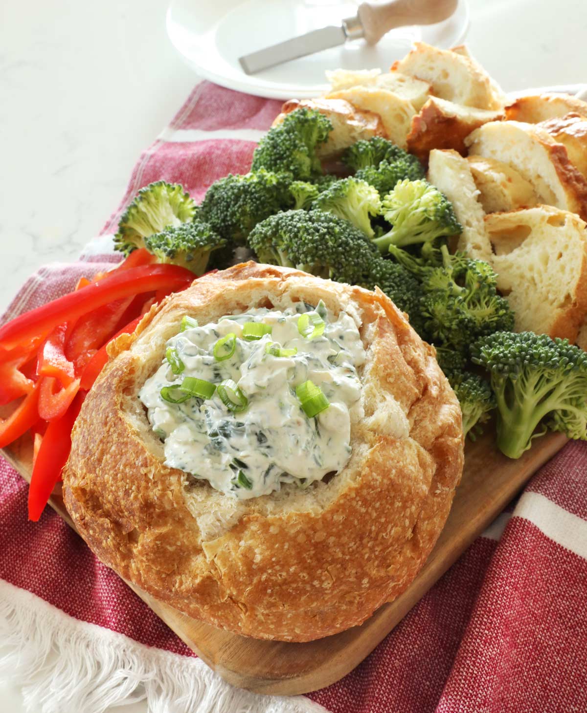 A close up of a bread bowl full of Knorr spinach dip sits on a wooden board along with some broccoli florets, sliced red bell pepper and more bread pieces, a cheese spreader sits on a white plate in the background on top of the kitchen counter.
