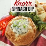 A close up of a bread bowl full of Knorr spinach dip sits on a wooden board along with some broccoli florets, sliced red bell pepper and more bread pieces.