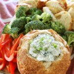 A bread bowl full of Knorr spinach dip sits on a wooden board along with some broccoli florets, sliced red bell pepper and more bread pieces, all on top of a red kitchen towel.