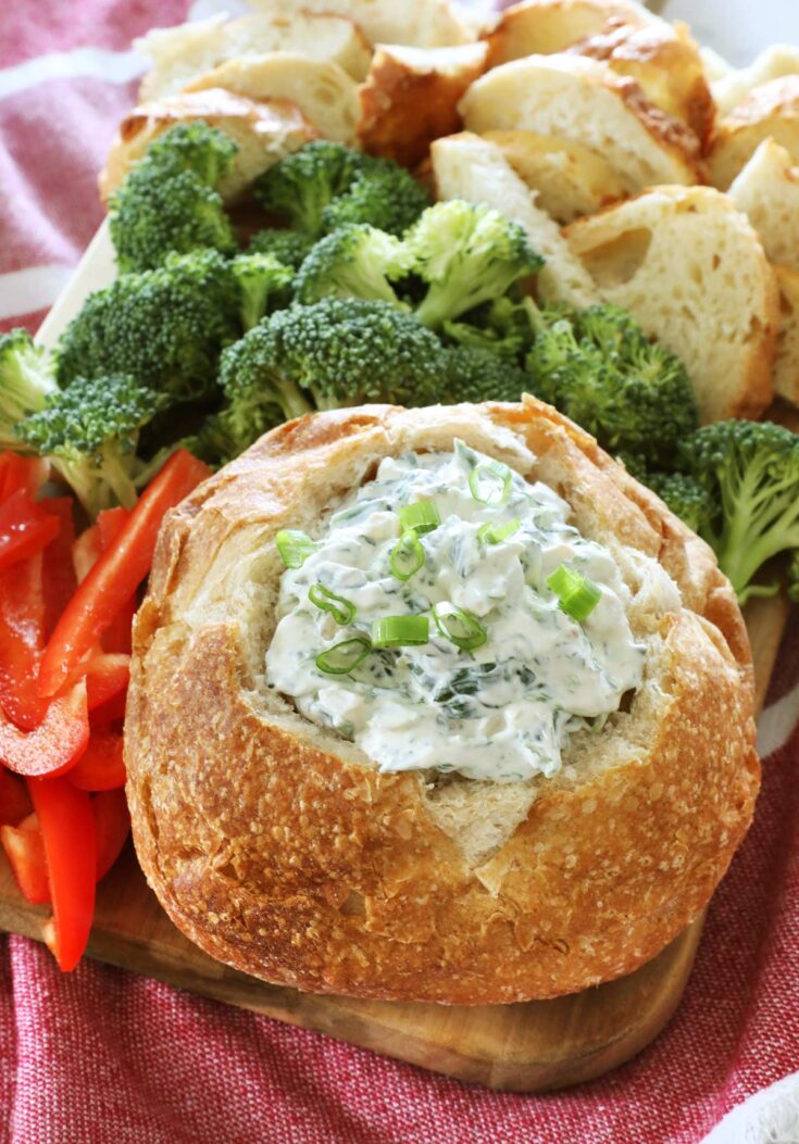 A close up of a bread bowl full of Knorr spinach dip sits on a wooden board along with some broccoli florets, sliced red bell pepper and more bread pieces, all on top of a red kitchen towel.