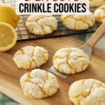 Four Lemon Crinkle Cookies sit on a wooden board, one actively being scooped up by a metal spatula while more lemon crinkle cookies cool on a rack in the background.