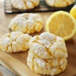 A stack of 3 lemon crinkle cookies are on a wooden board with more lemon crinkle cookies and a cut lemon in the background.