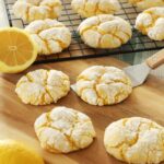 Four Lemon Crinkle Cookies sit on a wooden board, one actively being scooped up by a metal spatula while more lemon crinkle cookies cool on a rack in the background on the kitchen counter.