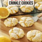 Four Lemon Crinkle Cookies sit on a wooden board, one actively being scooped up by a metal spatula while more lemon crinkle cookies cool on a rack in the background.