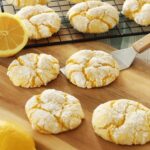 Four Lemon Crinkle Cookies sit on a wooden board, one actively being scooped up by a metal spatula while more lemon crinkle cookies cool on a rack in the background.