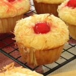 Piña colada muffin topped with a cherry on a wire cooling rack in a kitchen.